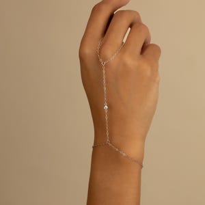 May include: A delicate silver hand chain with a small, clear gemstone. The chain connects to a ring and wraps around the wrist, creating a minimalist and elegant jewelry piece. The jewelry is designed to be worn on the hand.