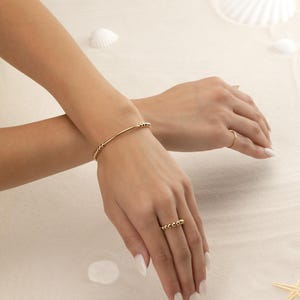 May include: A gold bracelet and ring set. The bracelet is a thin gold bar with small gold beads on each end. The ring is a thin gold band with small gold beads. The background is a sandy color with white seashells.