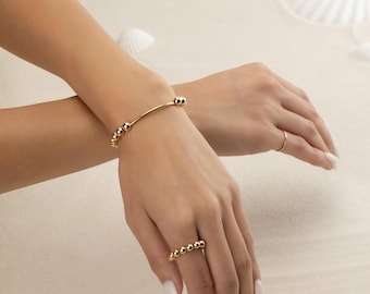 May include: Gold-toned jewelry set featuring a bracelet and a ring. The bracelet has a curved bar design with small, round beads. The ring also has a series of small, round beads. The jewelry is displayed on a light-colored surface.