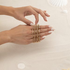 May include: A collection of gold bracelets, including beaded and chain styles, are displayed on a hand with manicured nails. The bracelets vary in design, with some featuring small beads and others larger, spherical beads. The background is a sandy texture.