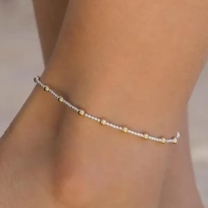 以下が含まれることがあります： チェーンに沿って均等に配置された小さな金色のビーズが付いた、繊細なシルバーのアンクレット。 アンクレットは足首に着用され、シンプルでありながらエレガントなデザインを際立たせています。 このジュエリーは、さりげない輝きを添えています。