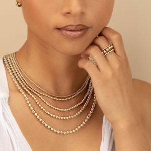 May include: A layered set of four gold bead necklaces of varying lengths, featuring small, polished spherical beads. The jewelry set includes a matching gold hoop earring and a stack of three thin gold bead rings worn on the finger.