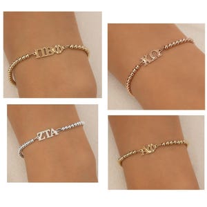 May include: Four beaded bracelets featuring Greek letter charms. The top left is gold with Pi Beta Phi, top right is gold with Chi Omega, bottom left is silver with Zeta Tau Alpha, and bottom right is gold with Alpha Phi letters.