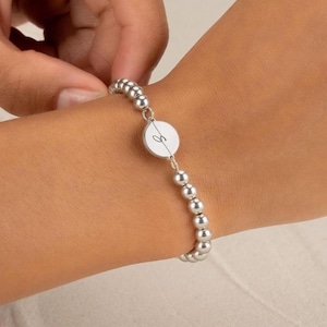 May include: A silver-toned beaded bracelet featuring a central circular charm. The charm is divided by a thin vertical line and displays a cursive letter S engraved on the surface. The bracelet is composed of small, uniform, polished metallic spheres.