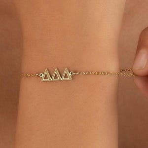 May include: A gold-colored bracelet featuring three connected triangle charms. The delicate chain is attached to the charms and is being held by a hand. The bracelet is a piece of jewelry.