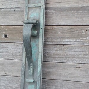 May include: A vintage, rectangular door handle made of metal with a patina finish. The handle is attached to a decorative plate with a rectangular opening. The handle is in the shape of a loop and is designed to be pulled.