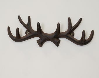 Könnte beinhalten: Ein brauner Wandhaken in Form eines Hirschgeweihs aus Gusseisen. Er ist an einer weißen Wand befestigt und hat mehrere Hängepunkte. Dieses rustikale Dekorationselement verleiht jedem Raum einen Hauch von Natur.