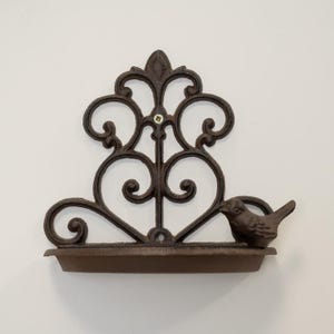 May include: A dark brown cast iron wall shelf with an ornate, scrolled design. A small bird is perched on the side. The shelf includes a small tray at the bottom. The shelf is designed for wall mounting.