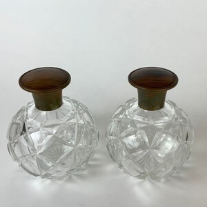 May include: Two clear glass bottles with a faceted design and brown wooden stoppers. The bottles are empty and have a vintage look.