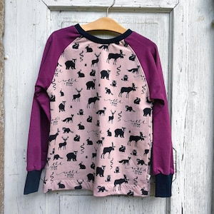Può includere: Una maglia a maniche lunghe con un pannello frontale rosa chiaro caratterizzato da un motivo nero ripetuto di orsi, cervi, alci, volpi e il testo "wild + free". Le maniche sono viola scuro con polsini e scollo di colore blu scuro.