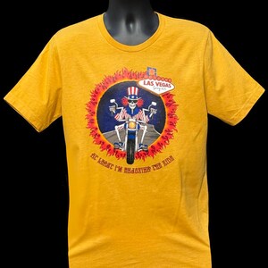 May include: Yellow t-shirt with a graphic of a skeleton riding a motorcycle in a circle of flames. The skeleton is wearing a red, white, and blue Uncle Sam hat and outfit. The text "At least I'm enjoying the ride" is below the graphic. The text "Las Vegas" is above the graphic.