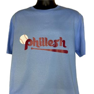 May include: A light blue t-shirt with a red baseball bat and the word "Phillesh" printed on the front. A white baseball is above the word.