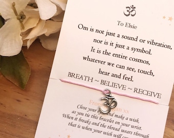 Peut inclure: Une carte blanche avec un cordon rose attaché autour. La carte a un charme en argent représentant le symbole Om attaché au cordon. La carte porte le texte "To Elsie" et une citation sur la signification d'Om. La carte porte également le texte "From ... gan xx" et "Naturally Bespoke".
