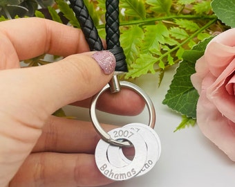 Puede incluir: Un llavero de cuero negro con un anillo de plata y dos dijes de plata. Los dijes están grabados con el texto "2007" y "Bahamas - tao".