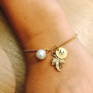 May include: A gold chain bracelet with a pearl charm, a gold leaf charm, and a gold disc charm with the letter "K" engraved on it.