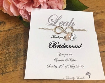 May include: A white card with the text "Leah Thank you for Bridesmaid Love you lots Lauren & Chris Sunday 26th of May 2019 Naturally Bespoke" and a silver infinity charm bracelet with a pearl and a letter L charm.