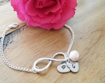 May include: A silver infinity bracelet with two heart charms engraved with the letters "G" and "C". A pink pearl charm hangs from the infinity symbol. The bracelet is lying on a wooden surface with a pink rose.