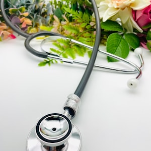 May include: A silver stethoscope with a black tube and a silver chest piece. The stethoscope is lying on a white surface with flowers in the background.