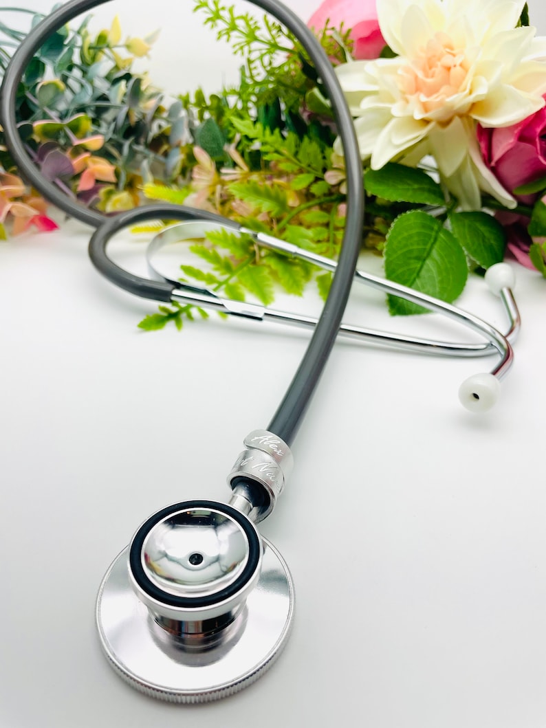 May include: A silver stethoscope with a black tube and a silver chest piece. The stethoscope is lying on a white surface with flowers in the background.