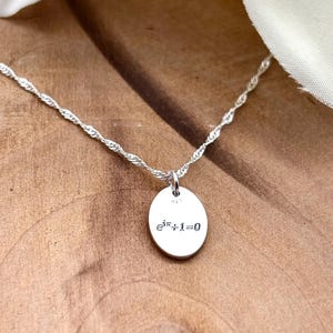 Op de afbeelding: Een zilveren ketting met een gedraaide ketting en een kleine, ovale hanger. De hanger is gegraveerd met de vergelijking e^πi+1=0. De ketting wordt gepresenteerd op een houten oppervlak, met een witte doek op de achtergrond.