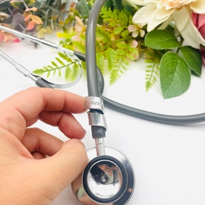 May include: A silver stethoscope with a gray tubing and a black rubber earpiece. The stethoscope is held in a hand and the tubing is coiled around the hand.