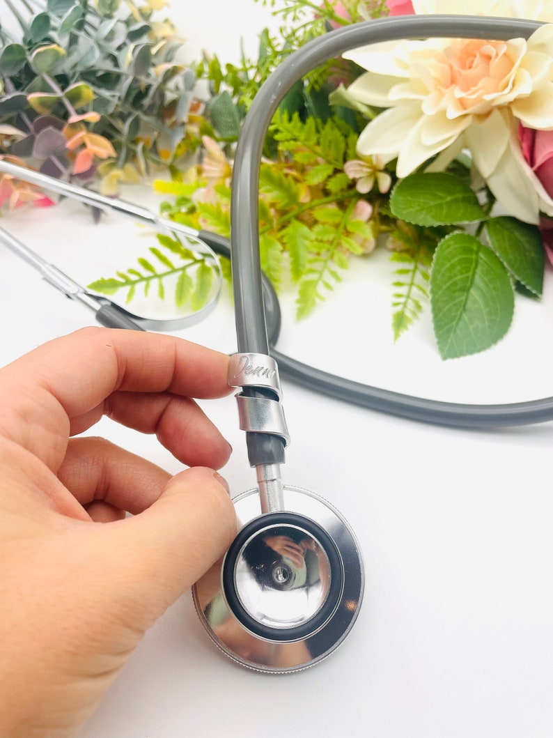 May include: A silver stethoscope with a gray tubing and a black rubber earpiece. The stethoscope is held in a hand and the tubing is coiled around the hand.