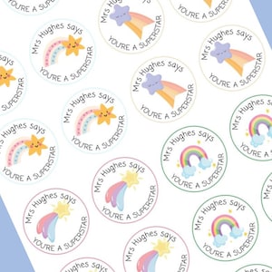 May include: A sheet of round stickers with a white background. Each sticker features a cartoon rainbow, star, or cloud with the text "Mrs Hughes says YOU'RE A SUPERSTAR" in a circular design. The stickers have pastel colors.