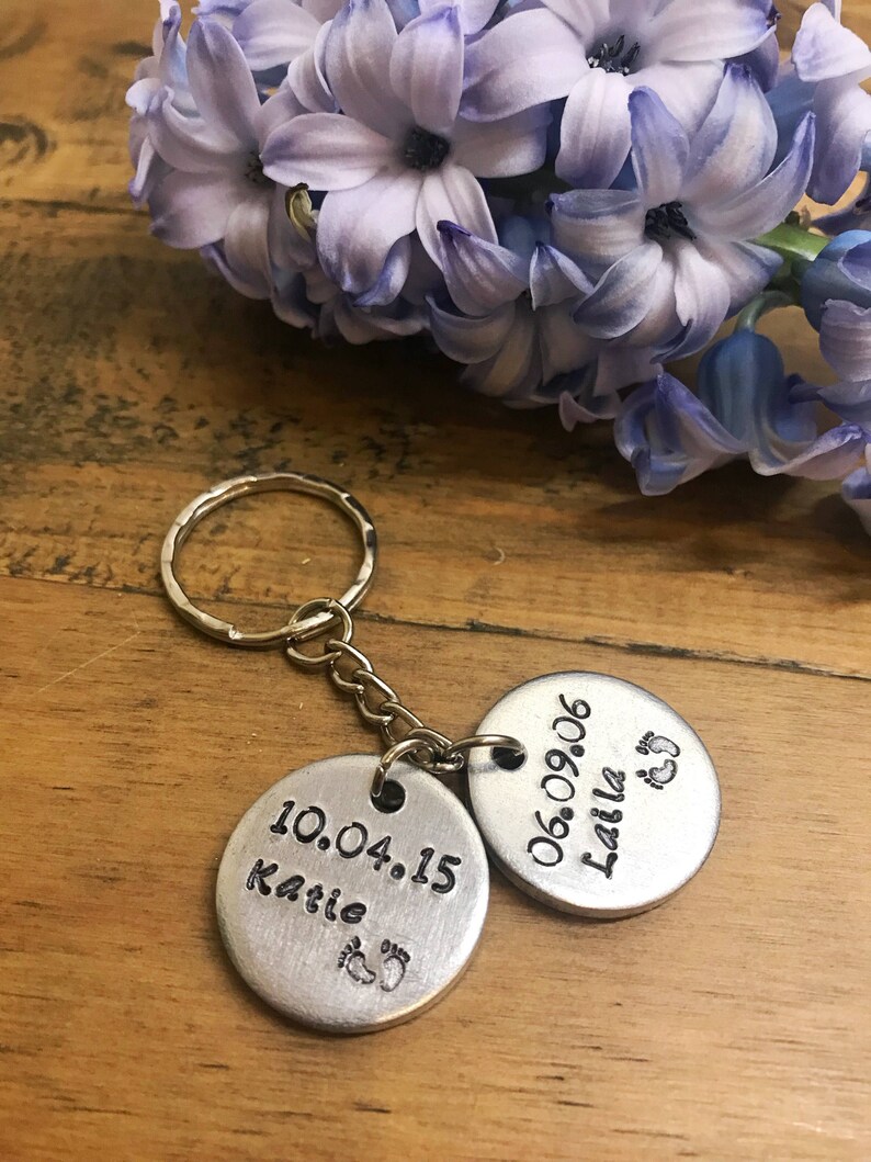 Peut inclure: Porte-cl&eacute;s argent&eacute; avec deux &eacute;tiquettes rondes en m&eacute;tal. L'&eacute;tiquette du haut indique "06.09.06 Laila" avec un symbole de pied. L'&eacute;tiquette du bas indique "10.04.15 Katie" avec un symbole de pied.