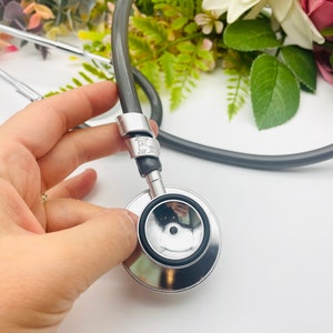 May include: A silver stethoscope with a black rubber tube and a silver earpiece. The stethoscope has a silver ring with the text "Joe 20" engraved on it.