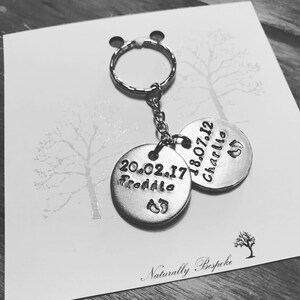 Peut inclure: Porte-cl&eacute;s en argent avec deux &eacute;tiquettes rondes. Une &eacute;tiquette dit "20.02.17 Freddie" avec un symbole de pied. L'autre &eacute;tiquette dit "18.07.12 Charlie" avec un symbole de pied. Le porte-cl&eacute;s est sur une carte blanche avec le texte "Naturally Bespoke".