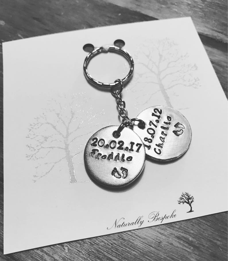 Peut inclure: Porte-cl&eacute;s en argent avec deux &eacute;tiquettes rondes. Une &eacute;tiquette dit "20.02.17 Freddie" avec un symbole de pied. L'autre &eacute;tiquette dit "18.07.12 Charlie" avec un symbole de pied. Le porte-cl&eacute;s est sur une carte blanche avec le texte "Naturally Bespoke".