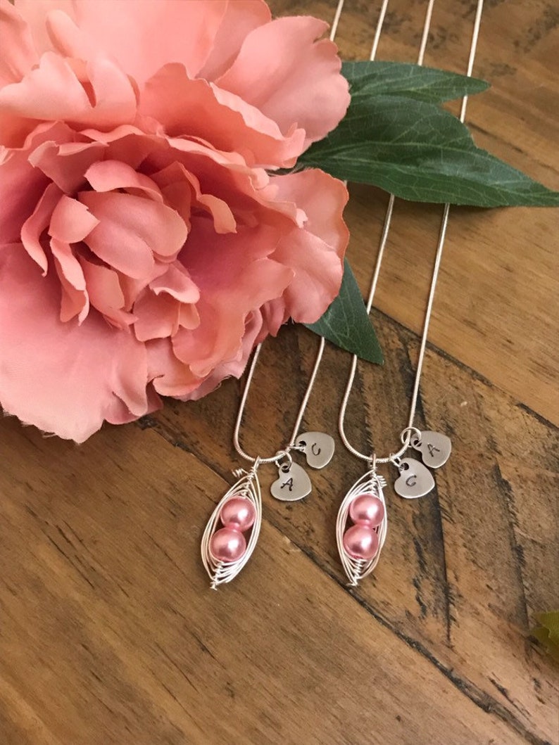 May include: Two silver necklaces with pink pearl pea pod charms and initial heart pendants. The necklaces are on a wooden surface with a pink flower.