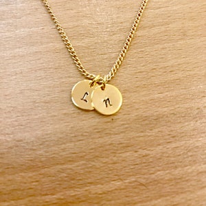 May include: A gold chain necklace with two small round gold charms. The charms have the letters "L" and "N" engraved on them.