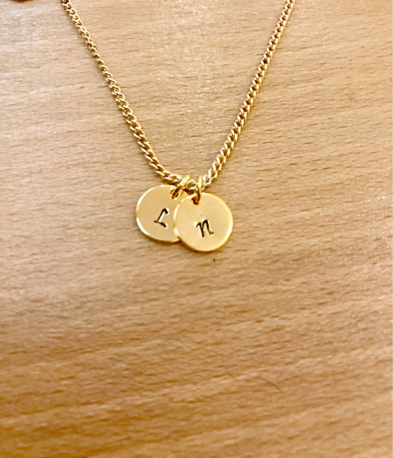 May include: A gold chain necklace with two small round gold charms. The charms have the letters "L" and "N" engraved on them.