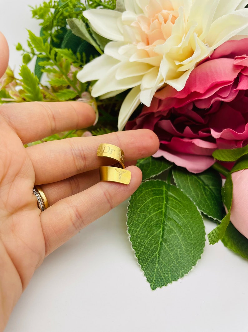 May include: Gold-toned ring with the inscription "Dr." and a medical symbol. The ring is shown on a finger.