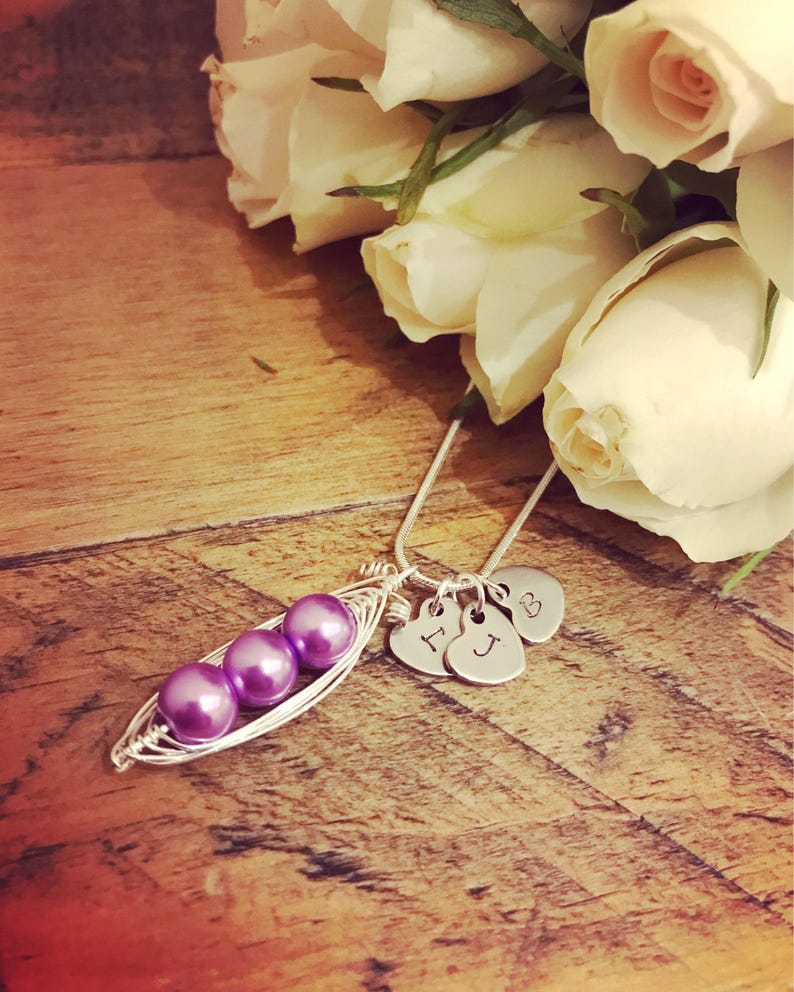May include: A silver necklace with a purple peapod charm and three silver heart charms with the initials I, J, and B. The necklace is lying on a wooden surface with white roses in the background.