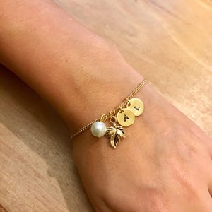 May include: A gold chain bracelet with three gold charms, each with a letter, A, L, and I. The bracelet also has a small white pearl charm and a gold leaf charm.