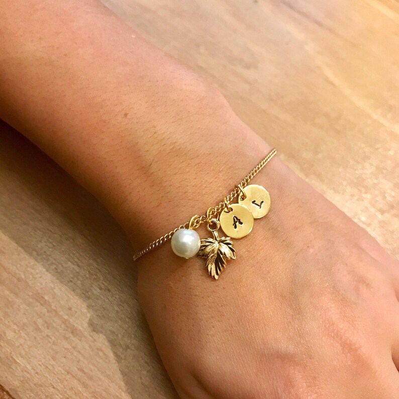 May include: A gold chain bracelet with three gold charms, each with a letter, A, L, and I. The bracelet also has a small white pearl charm and a gold leaf charm.