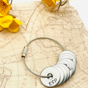 May include: A silver key chain with a series of round metal tags. The tags are engraved with letters, including the word "ESP". The keychain is lying on a vintage map with yellow flowers.