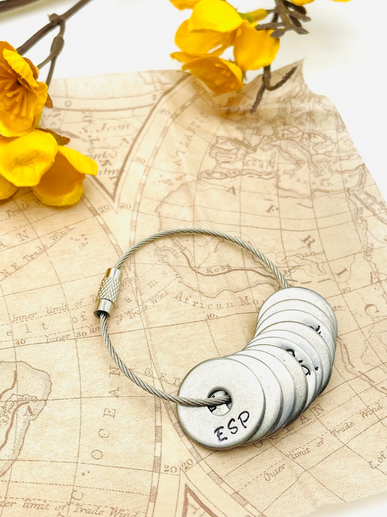May include: A silver key chain with a series of round metal tags. The tags are engraved with letters, including the word "ESP". The keychain is lying on a vintage map with yellow flowers.