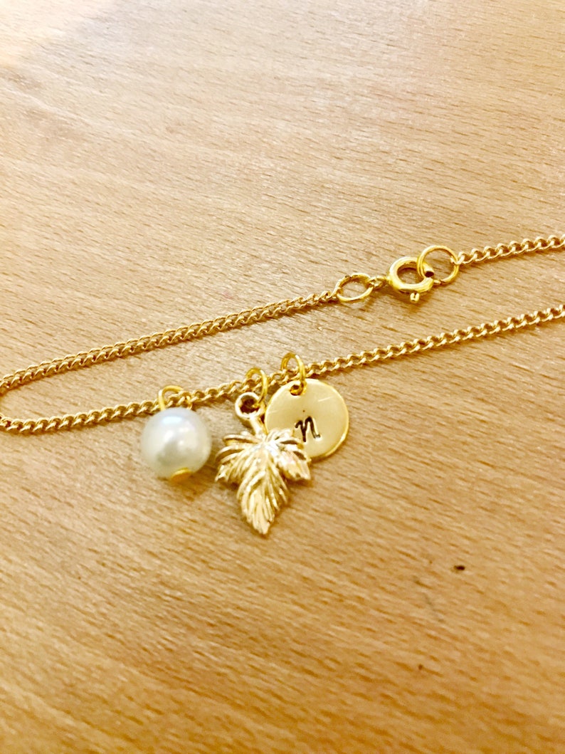 May include: A gold chain bracelet with a pearl charm, a gold leaf charm, and a gold disc charm with the letter "N" engraved on it.