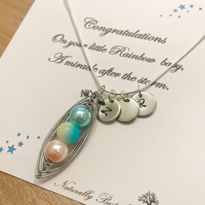 Puede incluir: Un collar de plata con un dije en forma de vaina de guisante con tres cuentas de color pastel. El dije est&aacute; unido a una cadena de plata con tres discos de plata que tienen las letras "M", "2" y "R" grabadas. El collar se muestra en una tarjeta blanca con el texto "Felicidades por tu peque&ntilde;o beb&eacute; Arco&iacute;ris, Un milagro despu&eacute;s de la tormenta. Naturalmente a medida".