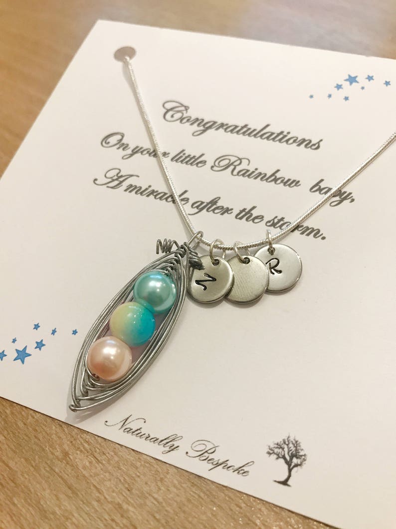 Puede incluir: Un collar de plata con un dije en forma de vaina de guisante con tres cuentas de color pastel. El dije est&aacute; unido a una cadena de plata con tres discos de plata que tienen las letras "M", "2" y "R" grabadas. El collar se muestra en una tarjeta blanca con el texto "Felicidades por tu peque&ntilde;o beb&eacute; Arco&iacute;ris, Un milagro despu&eacute;s de la tormenta. Naturalmente a medida".