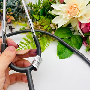 May include: A black stethoscope with a silver charm engraved with the names "Alex" and "Nurse".