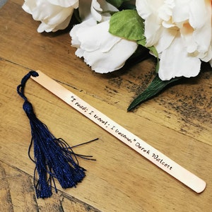 May include: A wooden bookmark with a blue tassel. The bookmark has the quote "I read; I travel; I become." by Derek Walcott engraved on it.