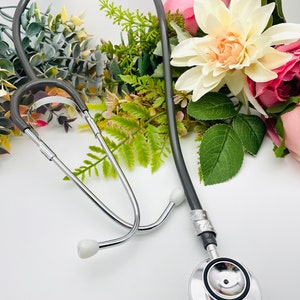 May include: A silver stethoscope with a gray tubing and a silver name tag. The stethoscope is lying on a white surface with a bouquet of pink and white flowers.