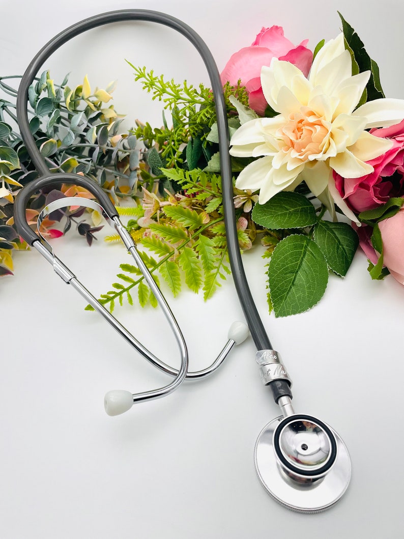 May include: A silver stethoscope with a gray tubing and a silver name tag. The stethoscope is lying on a white surface with a bouquet of pink and white flowers.