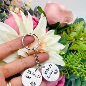 Peut inclure: Deux porte-cl&eacute;s en argent grav&eacute;s avec des dates et des noms. Le porte-cl&eacute;s de gauche indique "21.06.19 Ada" avec un symbole de pied. Le porte-cl&eacute;s de droite indique "30.04.21 Isaac" avec un symbole de pied.