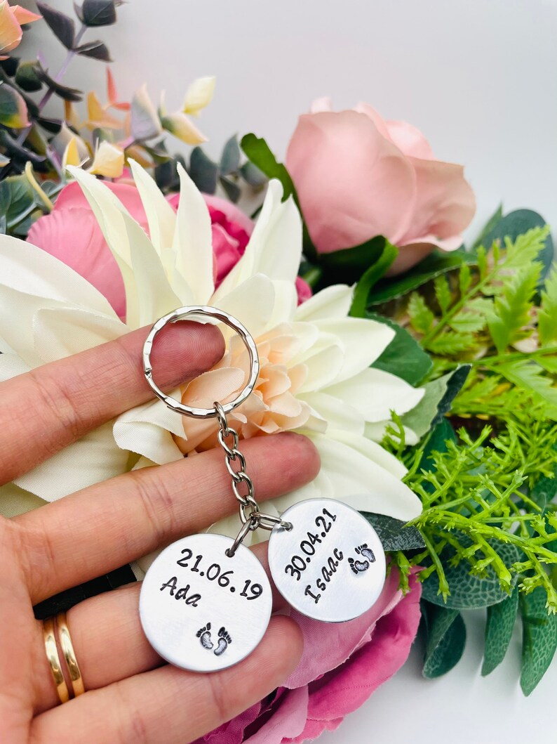 Peut inclure: Deux porte-cl&eacute;s en argent grav&eacute;s avec des dates et des noms. Le porte-cl&eacute;s de gauche indique "21.06.19 Ada" avec un symbole de pied. Le porte-cl&eacute;s de droite indique "30.04.21 Isaac" avec un symbole de pied.