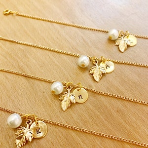 May include: Four gold chain bracelets with a pearl and a gold leaf charm with a letter on a round disc. The letters are K, N, A, and a fourth letter that is not visible.